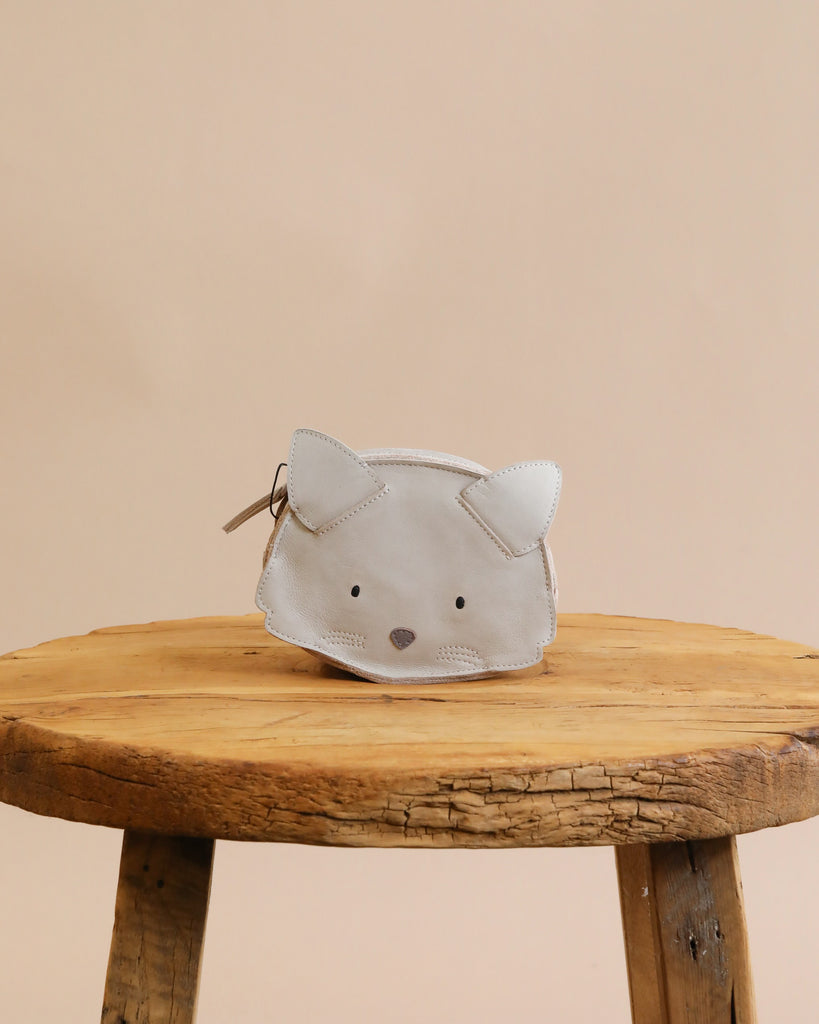A small Donsje Mini Leather Backpack - Cat designed to look like a cat's face, with stitched details and pointy ears, resting on a rustic wooden stool against a light beige background.