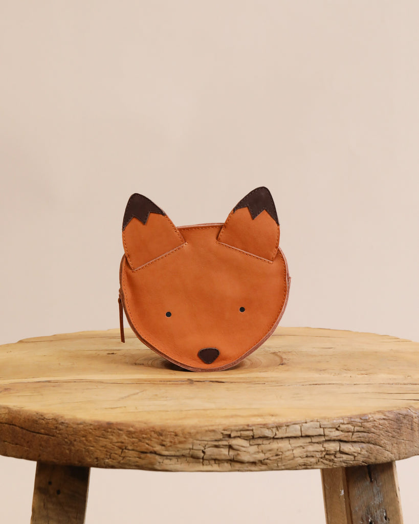 A Donsje Mini Leather Backpack - Fox rests on an old wooden stool against a plain beige background. The backpack features a simple face design with dark ear tips and is handmade fairtrade.