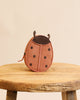 A round, 100% leather ladybug-themed Donsje Mini Leather Backpack with black spots, stitched details, and small antennae, displayed on a rustic wooden stool against a neutral background.