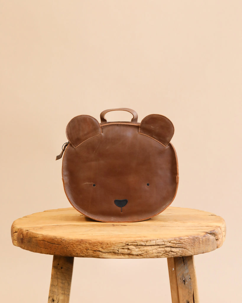 A Donsje School Leather Backpack - Bear designed to look like a bear's face, with small ears and features stitched in black, placed on a rustic wooden stool against a plain beige background.