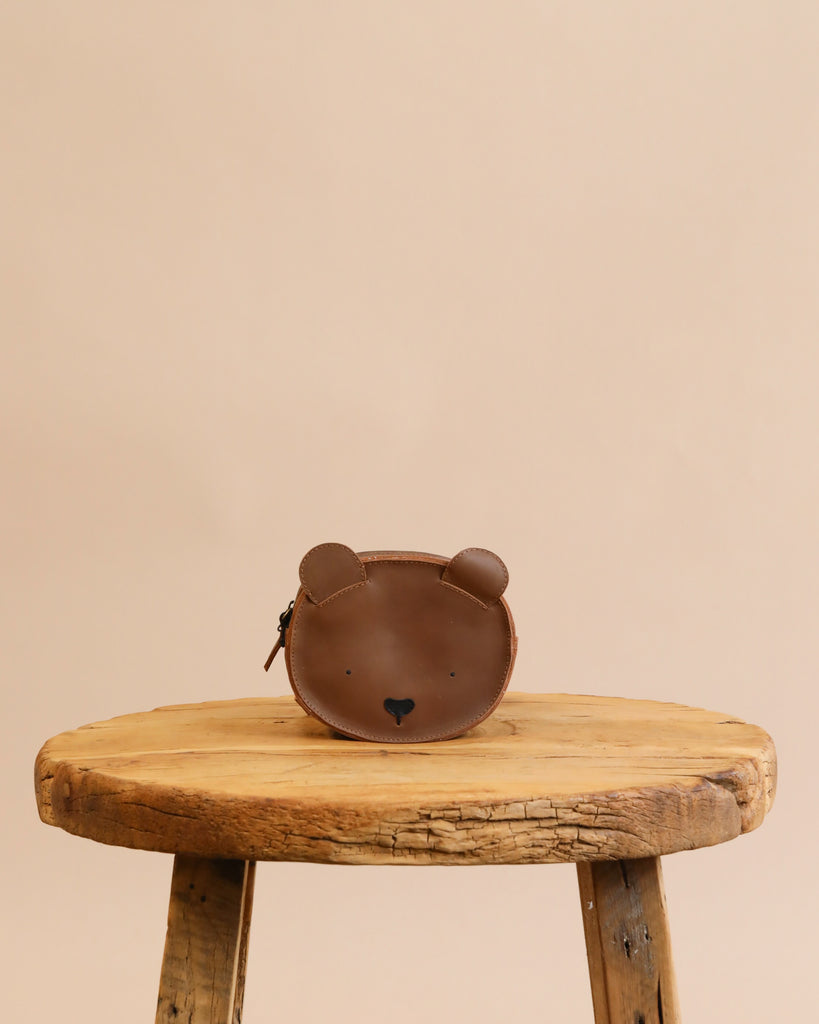A Donsje Mini Leather Backpack - Bear with a cute bear face design, including ears, on a rustic wooden stool against a plain beige background.