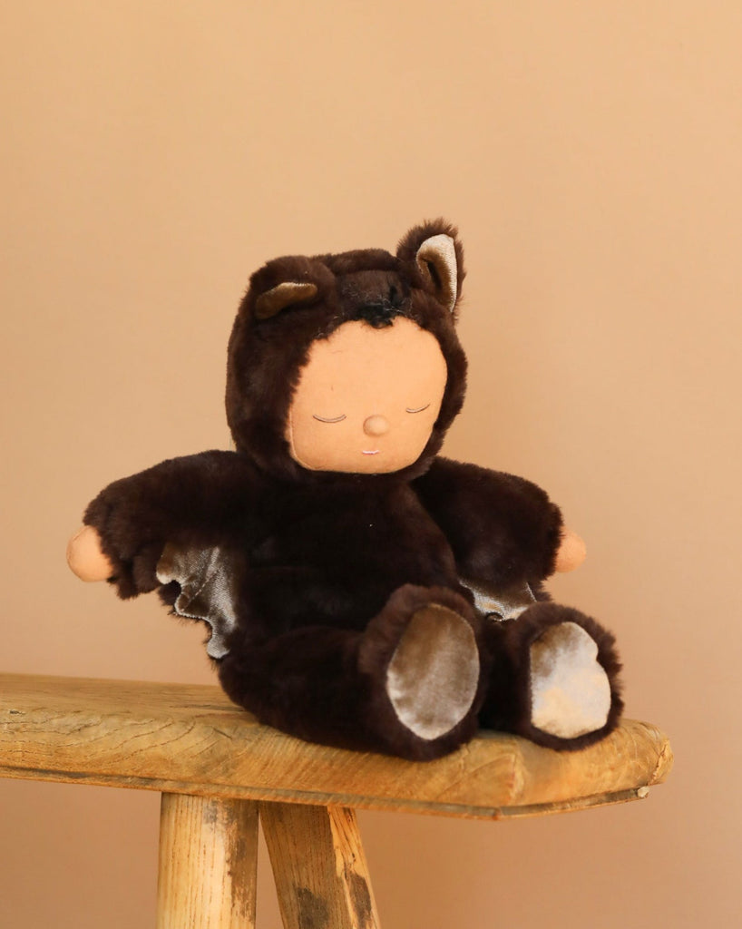 The Olli Ella Cozy Dinkum - Bat Ozzie - Dark Umber, a plush doll in a bat costume with ears, sits on a wooden stool against a beige background. This bedtime toy has a peaceful, sleeping face—perfect for cuddling.