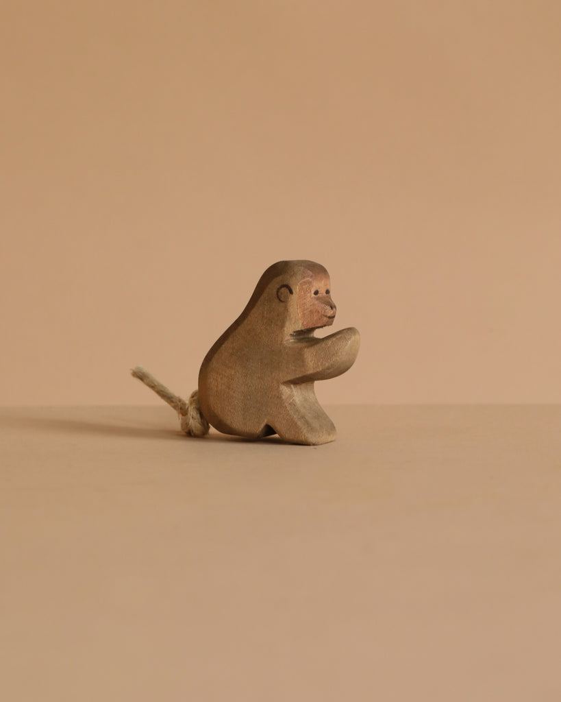 The Ostheimer Baboon, Sitting—a small, handcrafted wooden monkey with a rope tail—rests on a neutral beige surface against a matching background, capturing the iconic charm of Ostheimer wooden toys.