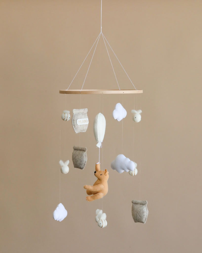 A baby nursery mobile featuring plush toys of a honey bear, clouds, and balloons, hanging from a wooden hoop against a beige background.