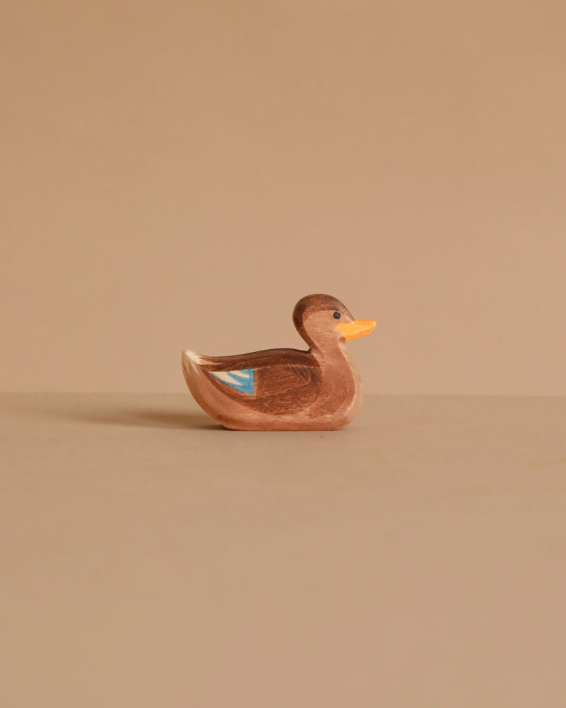 The Ostheimer Duck, Swimming is a small brown wooden figurine with a yellow beak and blue wing marking, handcrafted as a non-toxic toy and shown against a plain beige background.