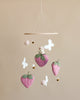 A nursery mobile featuring plush strawberries, white butterflies, and felt balls hanging from a wooden hoop against a beige background. A small figurine rests on one of the strawberries. Handmade Mobile - Field of Strawberries - Final Sale.