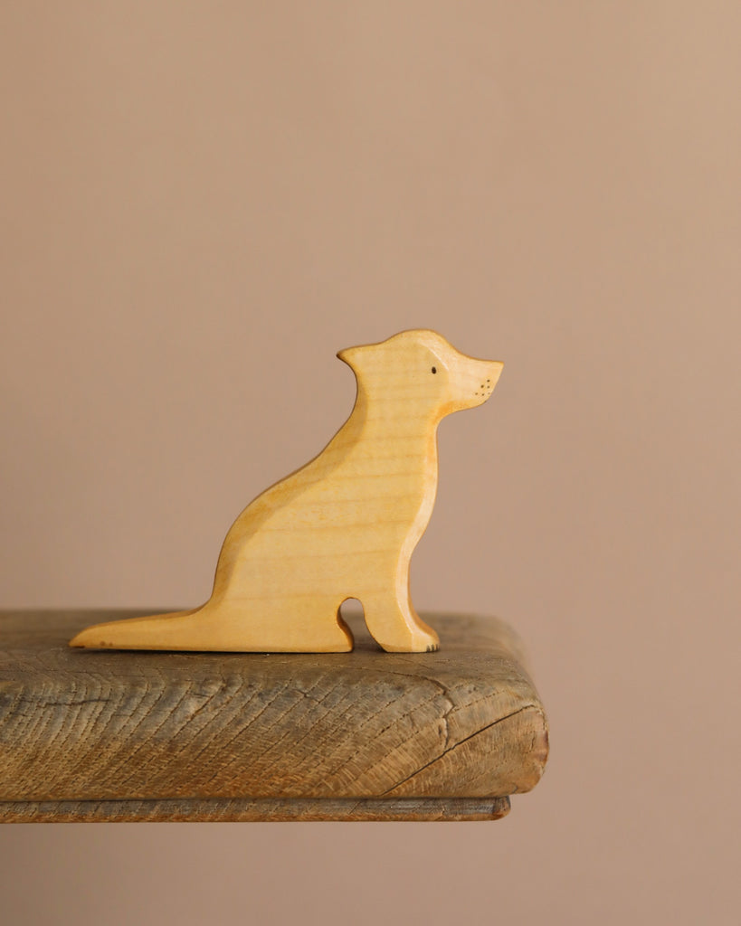A small Handmade Wooden Labrador Dog sits on a rustic wooden surface against a plain beige background. Crafted from sustainable hardwood, this light-colored figurine captures the essence of a sitting dog with simple, minimal features.