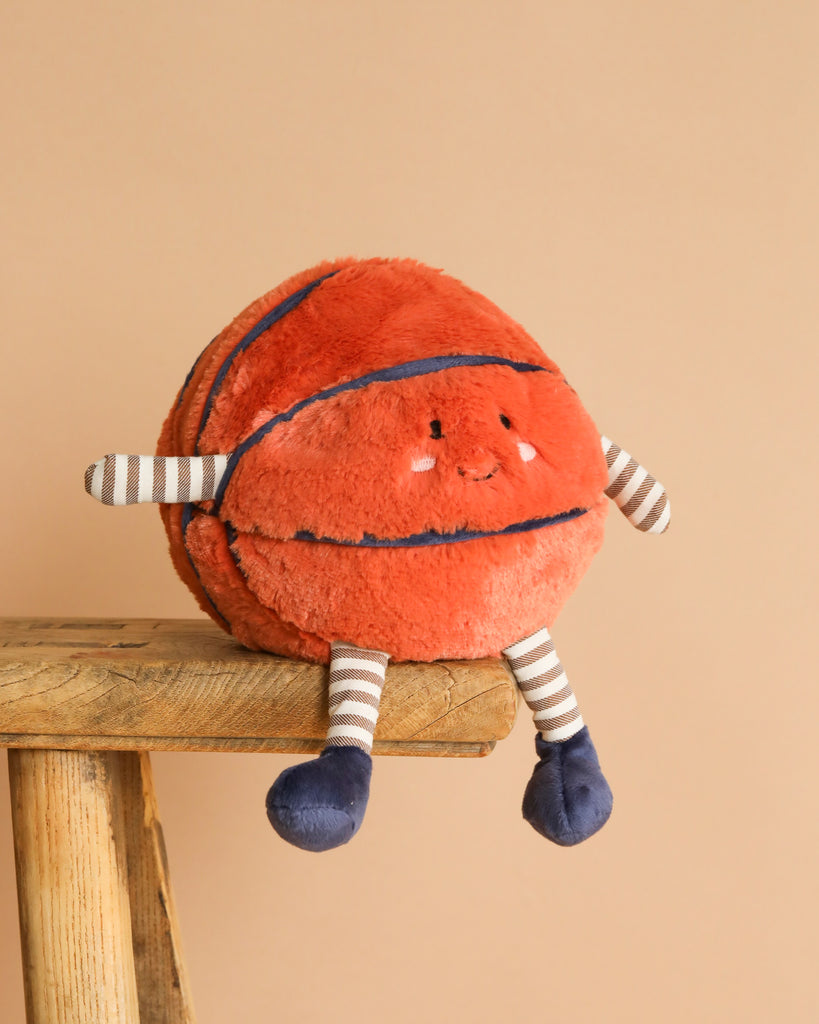 The Mon Ami - Barclay Basketball Plush Toy, featuring a smiling face, striped limbs, and dark blue feet, sits on a wooden stool against a beige background—making it an adorable baby gift.