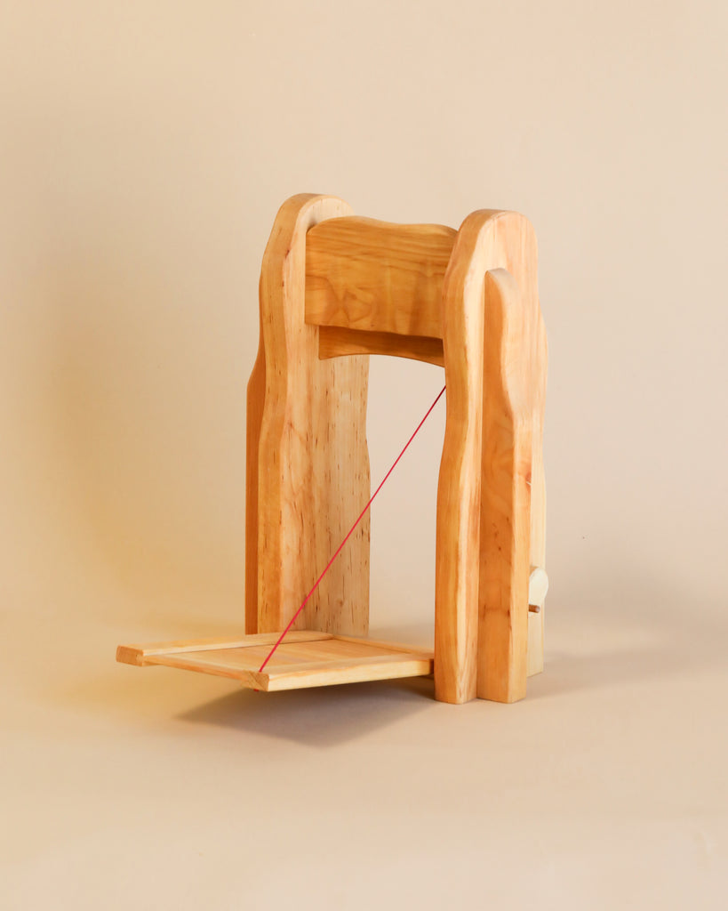 A wooden model reminiscent of Ostheimer Wooden Kingdom & Maileg Royal Family Set, featuring two side supports, a horizontal top beam, and a platform base held by red string. Set against a plain beige background.