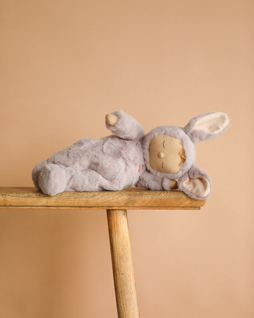 The Olli Ella Cozy Dinkum - Bunny Twinkle soft toy, dressed in a gray bunny costume with long ears and crafted from recycled plush for extra cuddles, lies on its side on a wooden bench against a beige background.