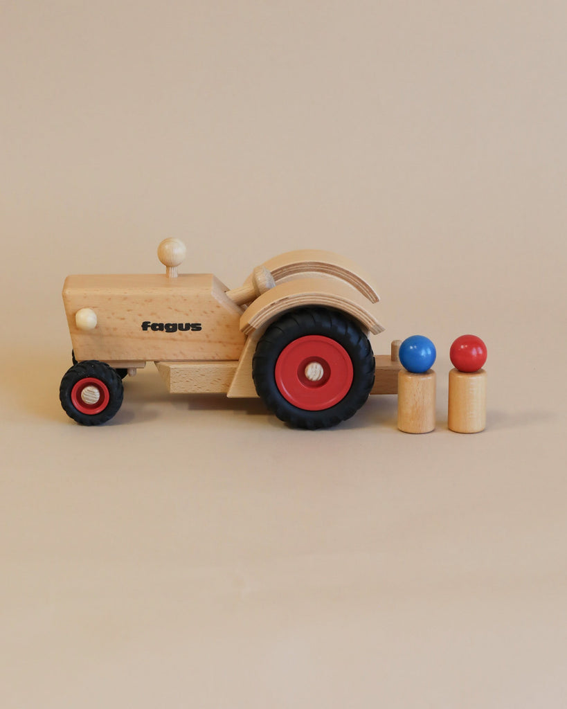A Fagus Wooden Tractor with the word "fagus" inscribed on the side, featuring red and black wheels, accompanied by three small peg figures in blue, red, and natural wood colors.