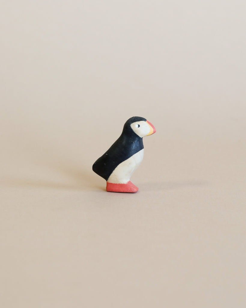 A small, colorful figurine of a puffin with a black and white body, handcrafted from linden wood, and a red base, standing upright on a plain beige background.