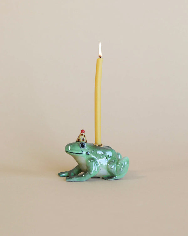 A hand-painted ceramic Frog Cake Topper with a lit yellow taper candle on its back, set against a neutral beige background. The frog has a playful expression and detailed green glazing.