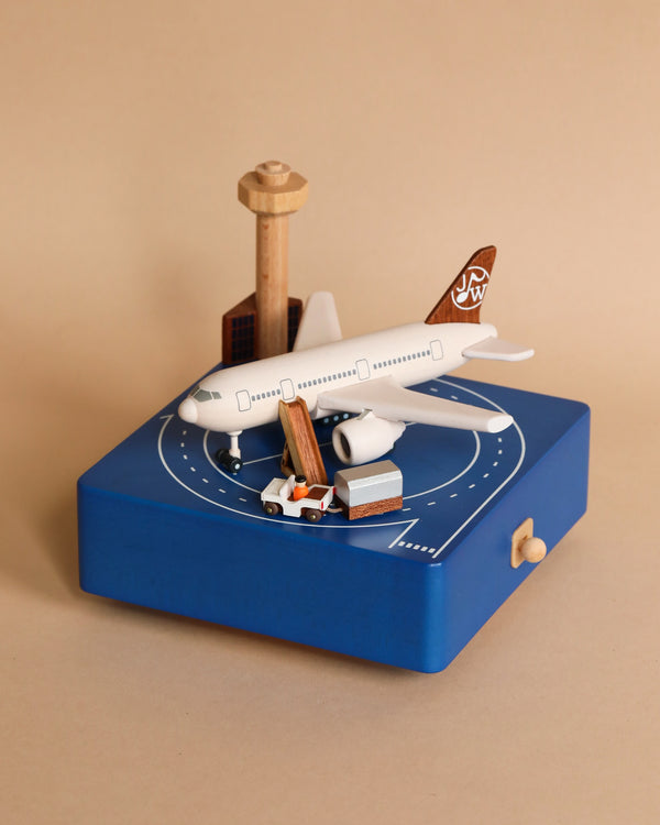 A miniature replica of an Airport Music Box sitting on a blue platform with a runway design, surrounded by small wooden models of buildings and vehicles, all crafted from sustainably sourced wood.
