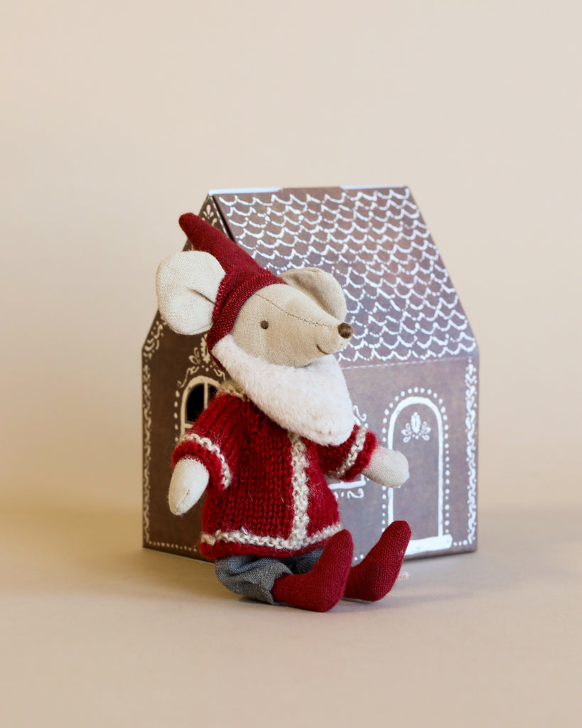 A festive Maileg Santa Mouse dressed in Santa clothes—a red hat, red sweater with white trim, gray pants, and red shoes—sits cheerfully in front of a Mouse Gingerbread House. The brown cardboard house adorned with white patterns evokes the charm of Christmas night. The background is beige.