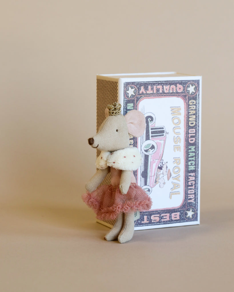 A Maileg Princess Little Sister mouse dressed in a pink tutu and a gold crown standing next to a vintage-style box labeled "mouse matchbox" in soft peach and pink colors, which is styled to resemble cozy bed linen.