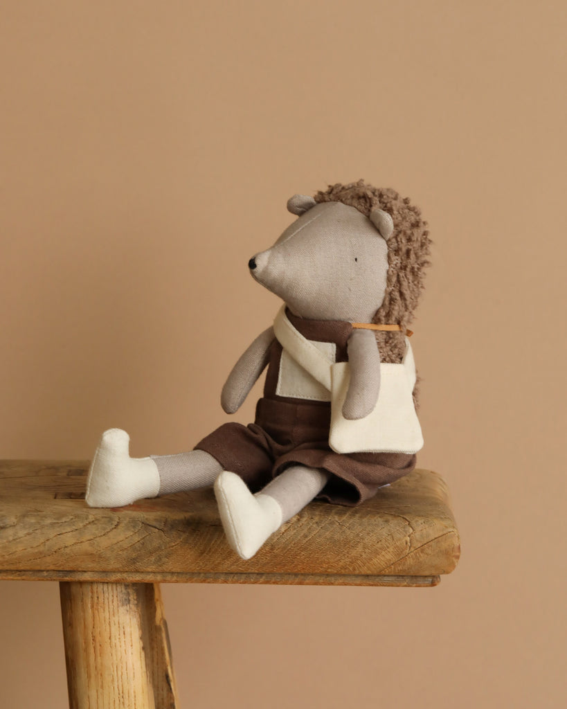 Harold The Hedgehog, part of the Hedgehog Family, sits on a wooden stool against a beige background. He wears a white top with brown overalls and has fluffy fur on his back. This cozy companion carries a small beige satchel, ready for secret garden adventures.