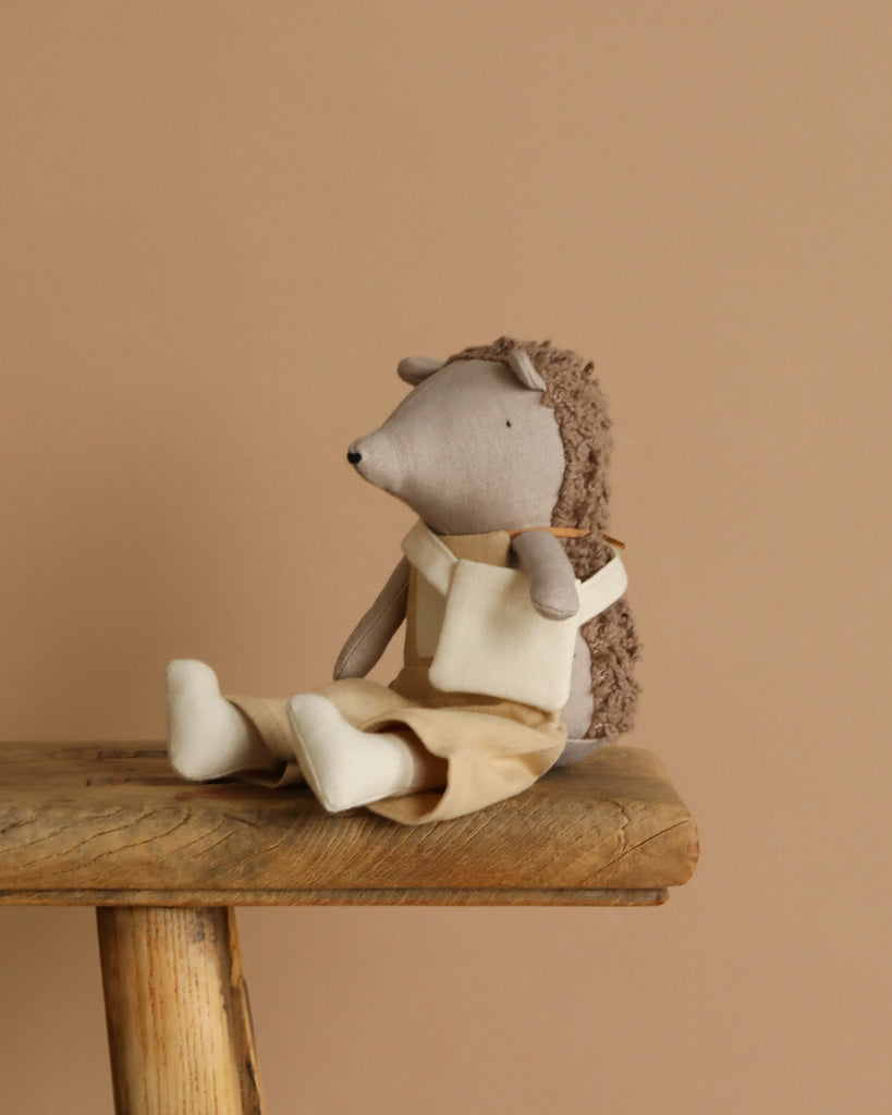 Heather the Hedgehog, a plush toy with enchanting charm, dons a beige outfit while perched on a wooden stool. The warm beige backdrop enhances the setting for her delightful garden escapades.