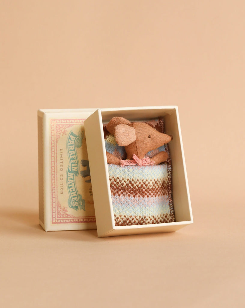 A small Maileg Sleepy Wakey Baby Mouse In Matchbox - Rose, tucked snugly inside a knitted blanket in a decorative box that resembles a matchbox.