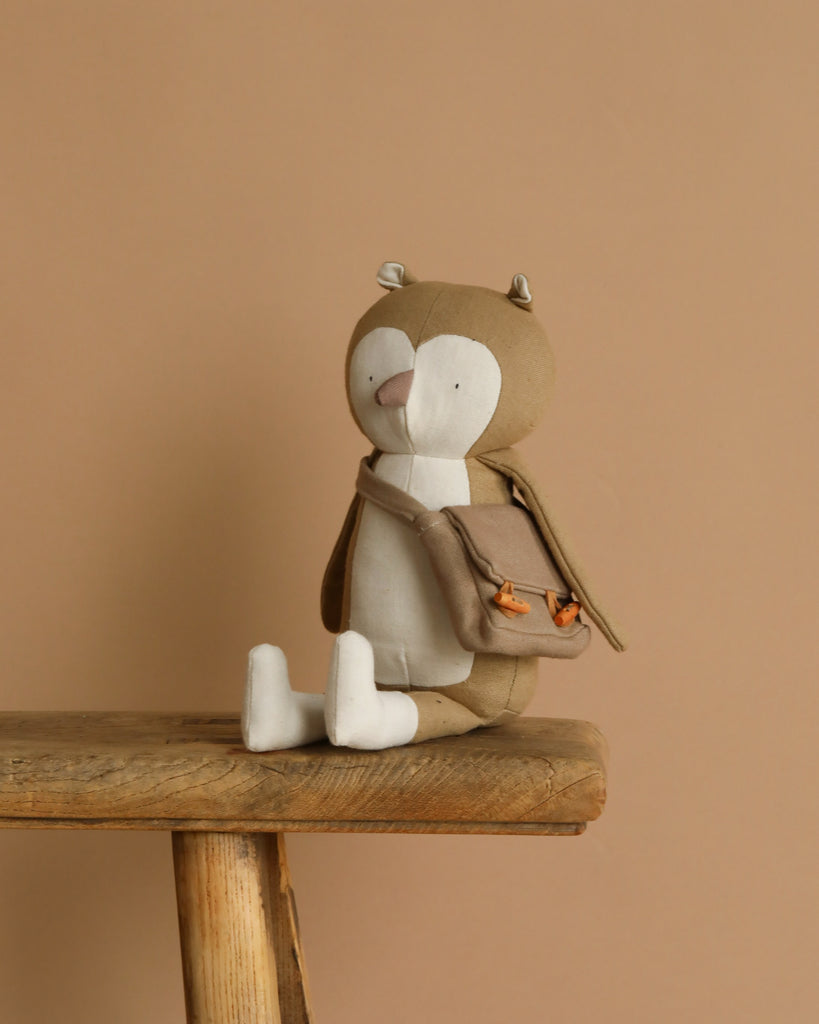 Owen The Owl, a woodland-themed plush toy in beige and white, perches on a wooden stool. Featuring small ears, a beak, and a tiny brown satchel, he seems set for an enchanting forest adventure against the smooth light brown backdrop.