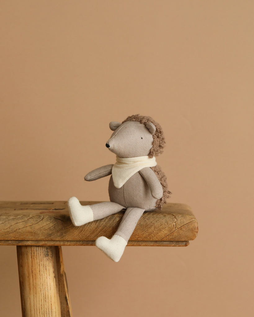 Poppet The Baby Hedgehog plush, wearing a light scarf and boots, sits on a wooden table. With its pocket-sized charm against a warm beige background, it creates an inviting and cozy atmosphere as an adorable garden companion.