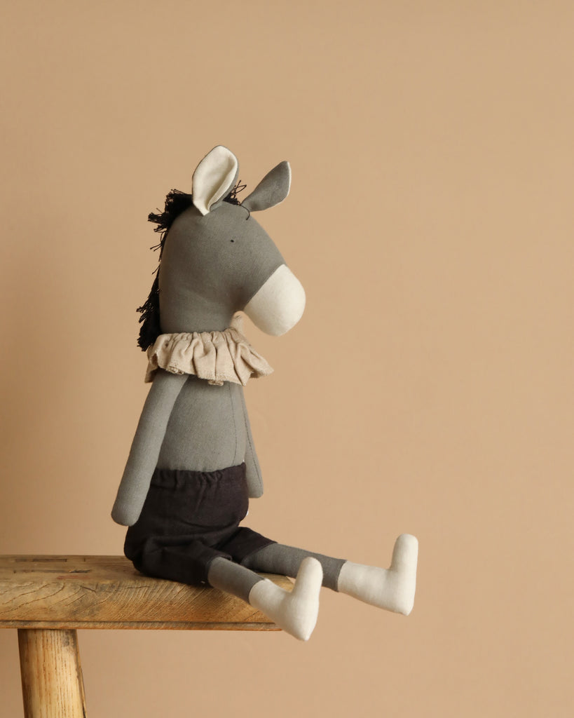 Meet Darwin the Donkey, a plush toy made from recycled polyester, featuring a white snout and ears. Dressed in a fluffy collar and dark shorts, it sits on a wooden stool against a beige background, with its legs charmingly dangling over the edge.