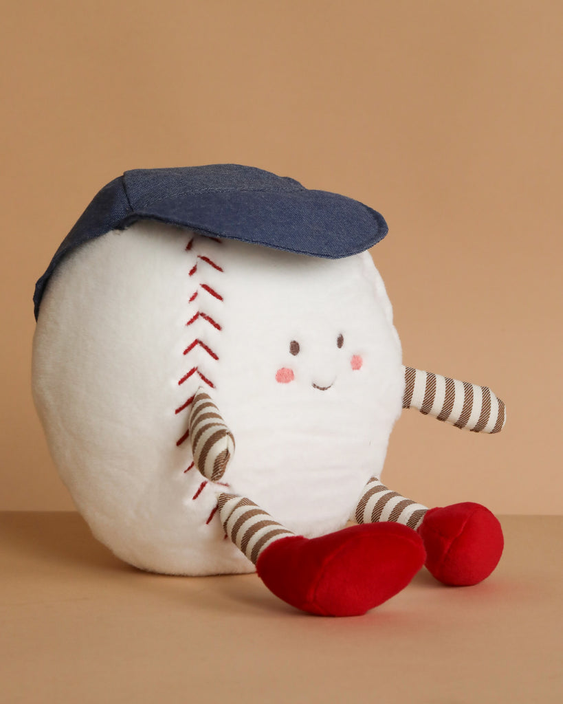 The Mon Ami - Buddy Baseball Plush Toy features stitched red seams, a blue cap, a smiling face, striped arms and legs with red feet—an adorable plush baseball perfect for cuddling or gifting, shown against a plain tan background.
