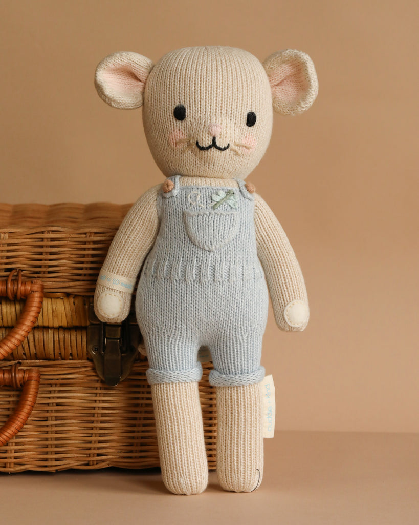 Cuddle + Kind Bailey The Mouse, a hand-knit doll in light blue overalls, stands before a wicker basket on a beige background and is crafted with hypoallergenic polyfill for gentle cuddles.