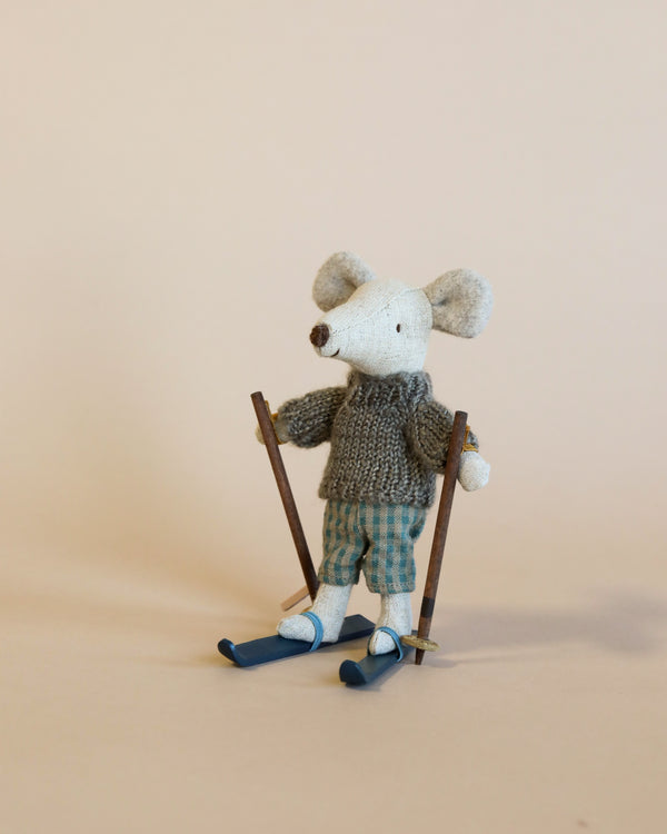 A small stuffed mouse toy, part of the Maileg Christmas Winter Mouse With Ski Set, Big Brother, stands upright wearing a gray knitted sweater and blue checkered pants. Holding two ski poles and with blue skis attached to its feet, it makes an adorable sight. Made from recycled polyester, the background is plain and light-colored.