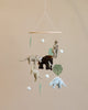 A nursery mobile featuring plush animals such as elephants and dinosaurs, accompanied by felt leaves and white orbs, hanging against a neutral backdrop.