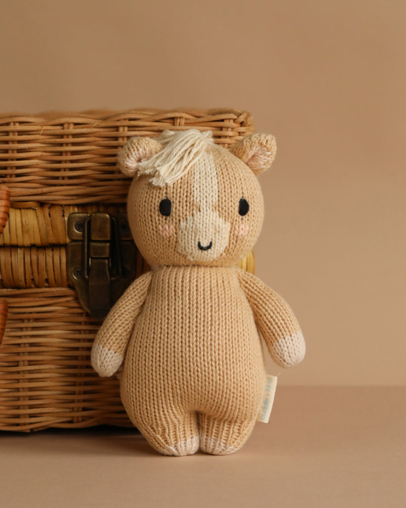 The Cuddle + Kind Baby Pony, a beige plush toy handmade in Peru from natural cotton yarn, features a smiling face, small ears, and a white tuft of hair. It stands by a wicker basket against a light brown background.
