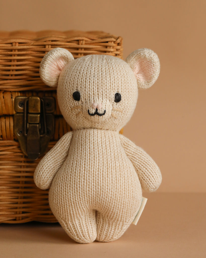 A beige, hand-knit Cuddle + Kind Baby Mouse with black eyes and a stitched smile sits upright by a woven wicker basket, crafted from natural Peruvian cotton yarn on a light brown surface.