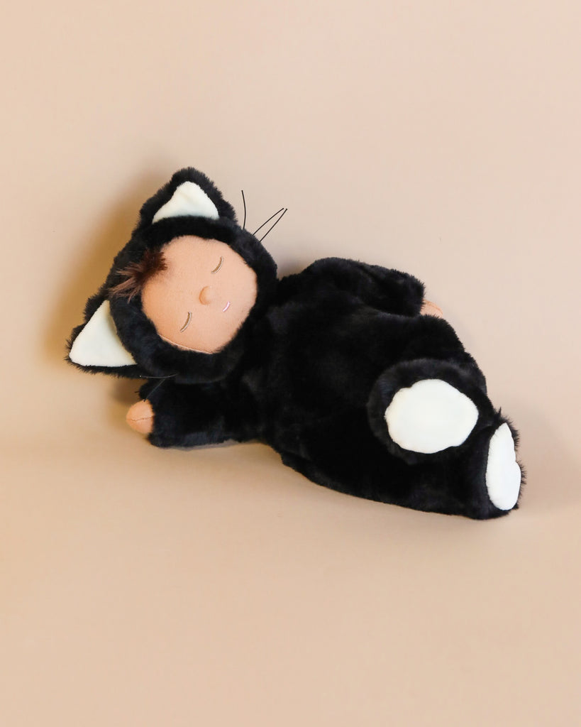 A plush toy resembling a black and white cat from the Olli Ella Cozy Dinkum Doll - Black Cat Nox collection with closed eyes, lying on its back on a pale pink background.