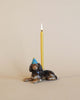 A Dachshund Cake Topper of a black dog wearing a blue party hat, with a lit candle inserted on top, set against a beige background.