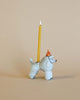 A whimsical Poodle Cake Topper with a yellow candle lit on its back, set against a plain beige background. The poodle wears a small, festive red hat with gold trim.
