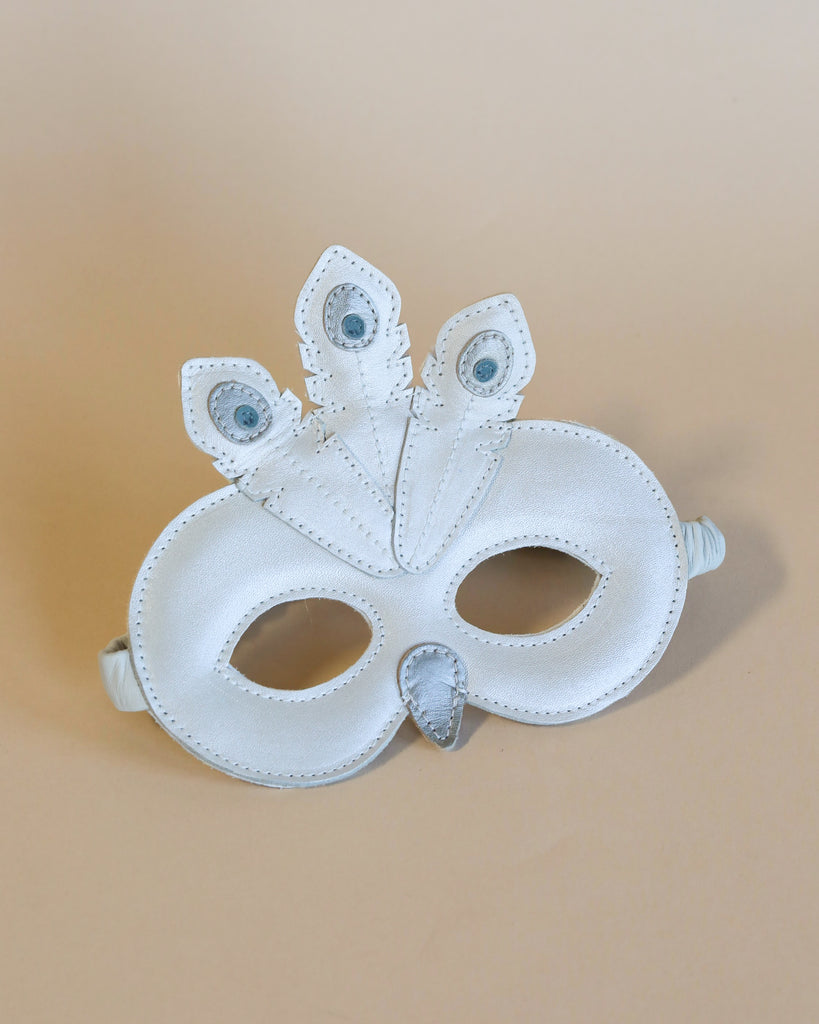 A white carnival mask with intricate designs and gemstone details, featuring three eye-like embellishments, against a soft beige background. This Donsje Tieri Mask | Peacock is adorned with metallic leather accents.