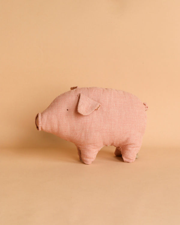 A close-up photo of a Maileg Polly Pork, Small, crafted from soft, pink plaid fabric and recycled polyester, standing on a light brown surface with a matching plain background. The toy features floppy ears and a small snout, giving it a charming handmade appearance.