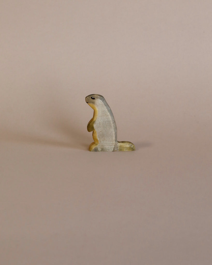 A small, solitary high-quality Handmade Holzwald Marmot figurine standing upright against a plain, light beige background. The figurine is textured and painted in natural colors.