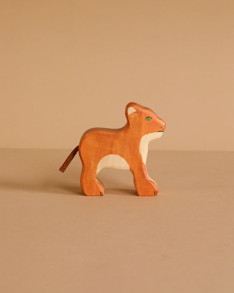 The Holztiger Lion Cub is a handcrafted wooden figure with a light brown body, cream face and chest, green eyes, and a short tail, displayed on a plain beige surface with a matching background.