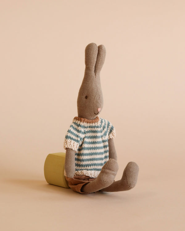 A soft natural linen rabbit sits upright against a green cylindrical object on a light beige background. The bunny, likely from a Maileg dollhouse, is dressed in a blue and white striped knit sweater with brown shorts. It has long ears and a simple, stitched facial expression. This is the Maileg Rabbit Size 1 - Brown - Shirt And Shorts.