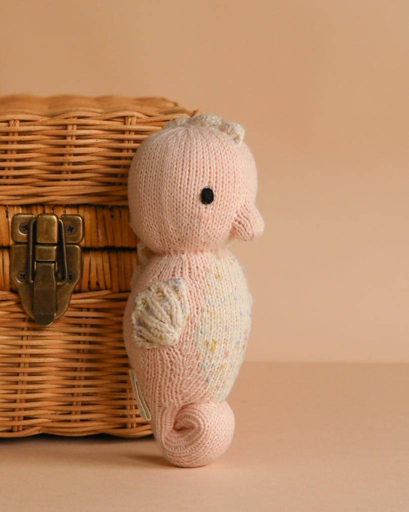The Cuddle + Kind Baby Seahorse in Powder Pink, hand-knit from heirloom-quality Peruvian cotton yarn, features a small fin and curled tail, resting beside a woven wicker basket with a brass latch on a peach background.