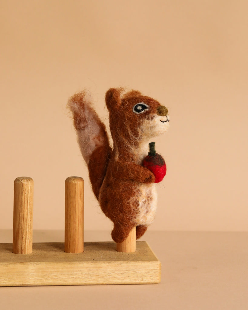 The Squirrel Finger Puppet, handcrafted by artisans in Nepal, features a brown felted squirrel standing upright and holding a small red object on a wooden base with three pegs, shown against a plain beige background.