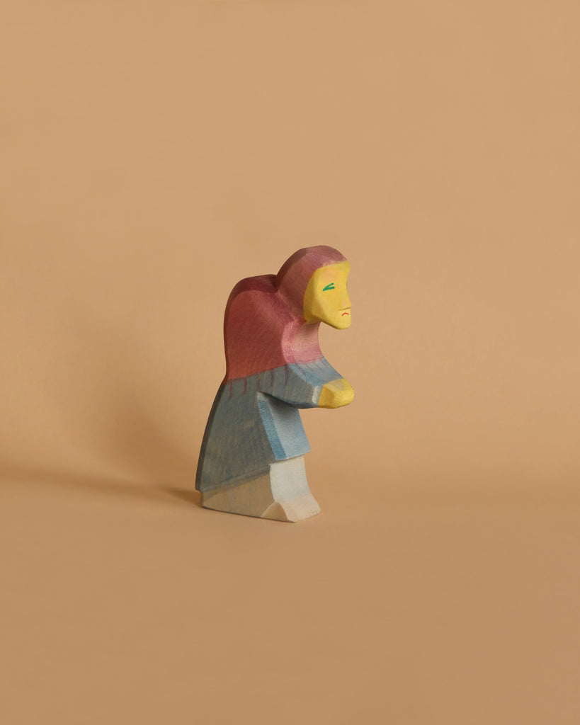 An Ostheimer Witch Set handcrafted wooden figurine, painted in simple colors, stands against a plain beige background. The figurine has a red and purple headscarf, a greenish blue top, and a cream-colored bottom half.