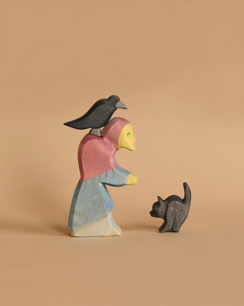 The Ostheimer Witch Set features a handcrafted wooden figure of a person wearing a pink headscarf and blue dress, made from sustainably sourced materials, standing on a beige background. A black bird perches atop the figure's head, while a small black cat with green eyes stands in front of them, gazing upward.