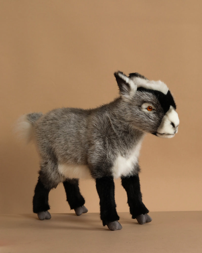 The Grey Goat Stuffed Animal features gray and white fur, black facial and leg markings, and lifelike yellow eyes. It stands on a light brown surface with a beige background, showcasing its realistic artisan craftsmanship.