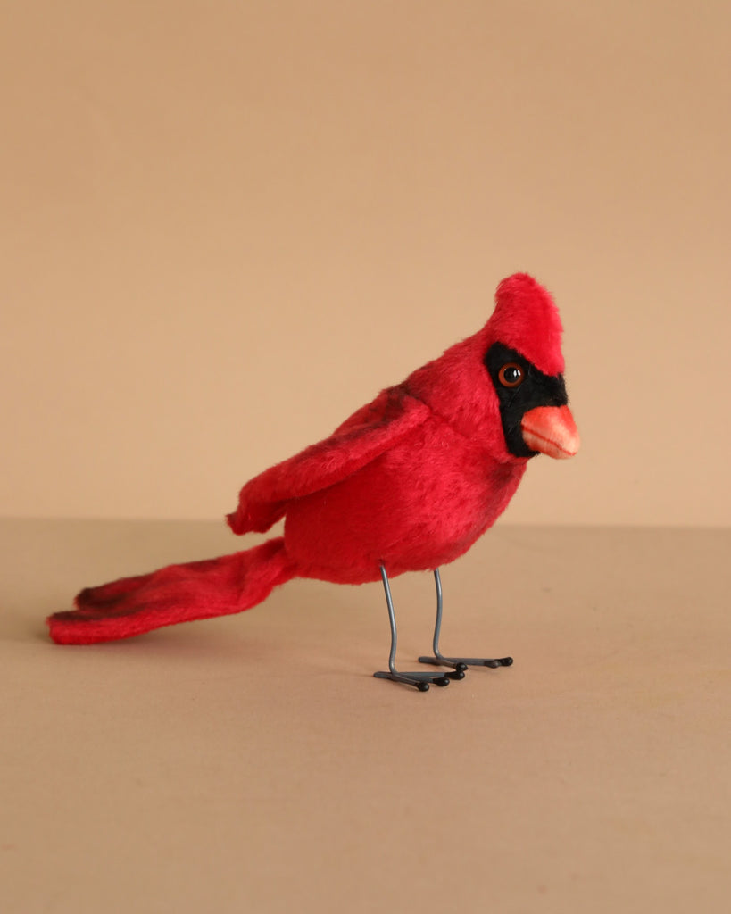 The Cardinal Red Bird features black facial markings and stands on thin wire legs, resembling a realistic plush animal, set against a plain beige background.