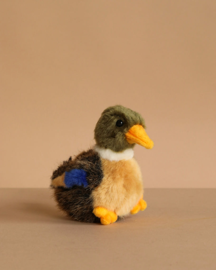 The Baby Duck Stuffed Animal is a small, plush toy with brown, green, and yellow fur, orange beak and feet, plus a blue patch on its wing—artisan crafted and shown against a plain beige background.