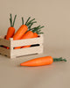 A wooden box filled with Erzi Carrot Pretend Food, specifically fresh orange carrots with green tops, handcrafted in Germany, placed on a neutral beige background. One carrot lies outside the box in the foreground.