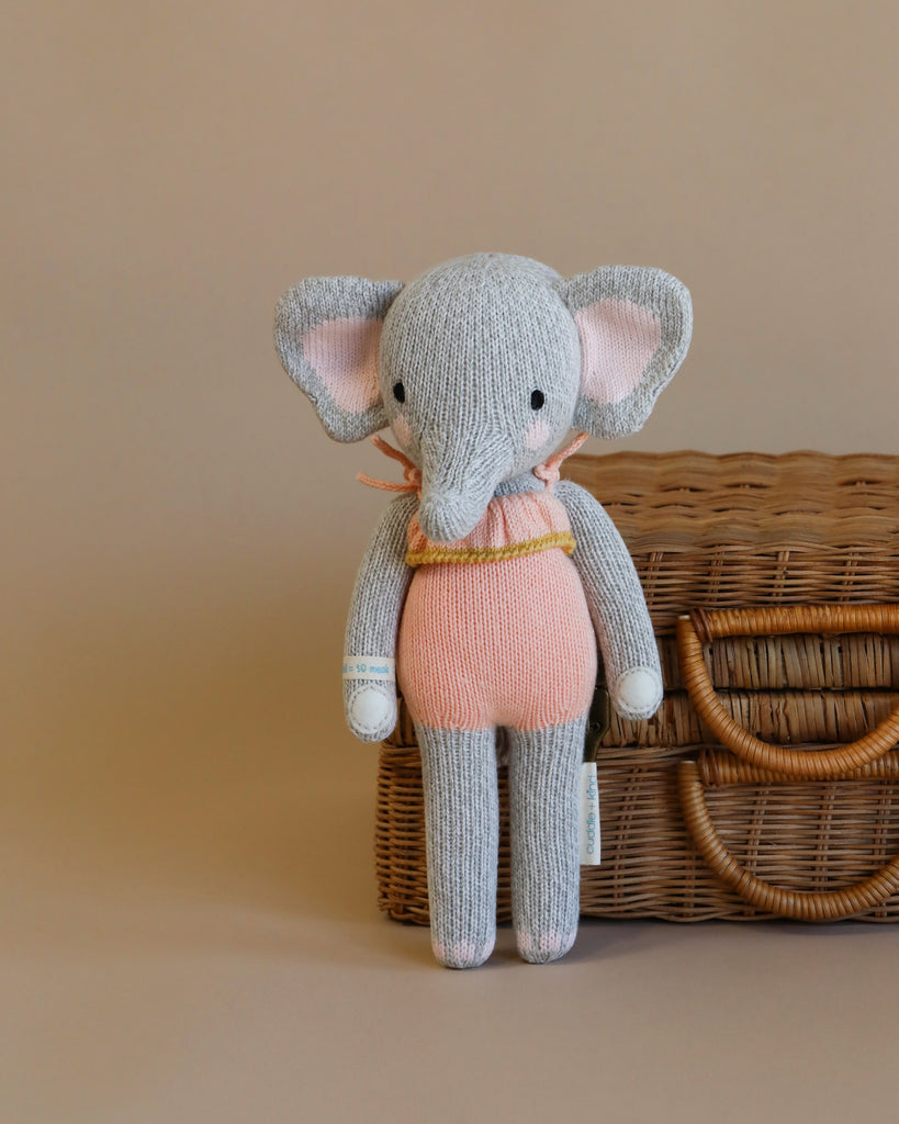 Cuddle + Kind Eloise the Elephant, a hand-knit gray stuffed doll with large ears, pink outfit, and yellow scarf, stands before a woven wicker suitcase on a beige background.