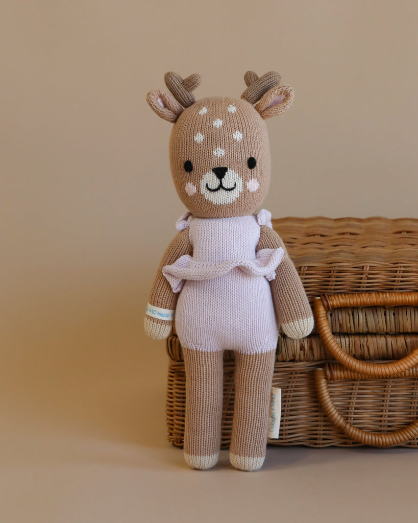 Cuddle + Kind Violet the Fawn is a hand-knit plush deer, made from premium cotton yarn, featuring cream forehead spots and a pink outfit, posed in front of a closed wicker suitcase on a beige background.